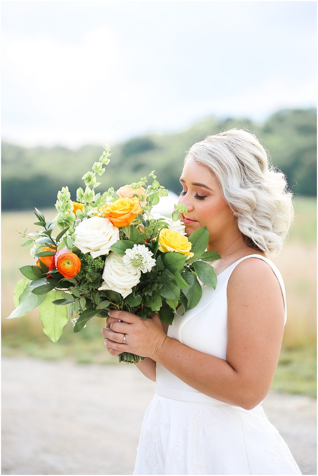 BRIDE SMELLING HER COLORFUL WEDDING BOUQUET FILLED WITH WHITE ORANGE AND YELLOW AT THE LEGACY AT GREEN HILLS FOR A WEDDING IN KANSAS CITY THAT WAS PHOTOGRAPHED BY MARIAM SAIFAN PHOTOGRAPHY - BEAUTIFUL WEDDING BOUQUET - KC WEDDING FLORIST + PHOTOGRAPHER