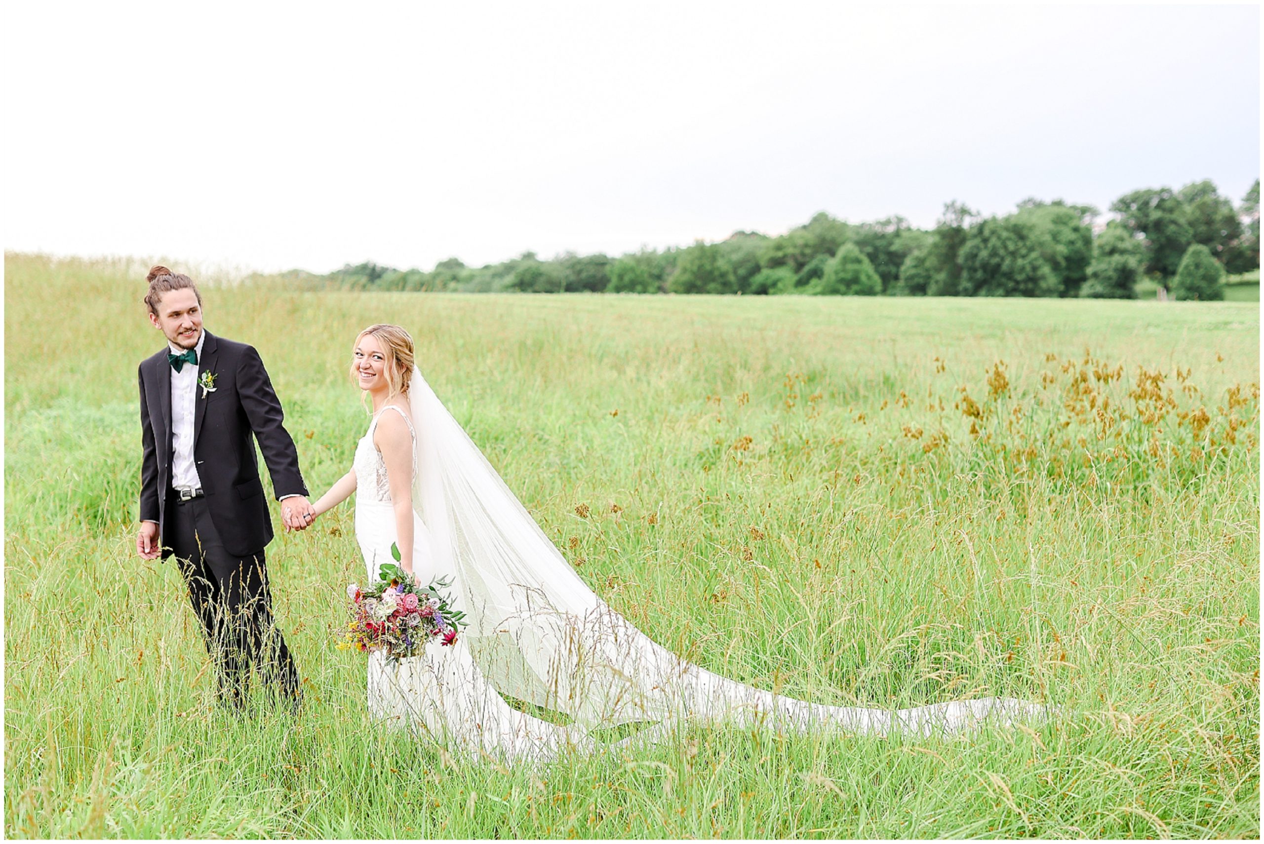 bride and groom poses walking in a field of tall grass - Powell Gardens - Wedding Venue in Kansas Missouri - Wedding Photography by Mariam Saifan Photography - Brianna & Kendall Summer Wedding - Emerald Wedding Colors - Best Engagement Photographer in Kansas City & Family Photos - Wedding Photography KC